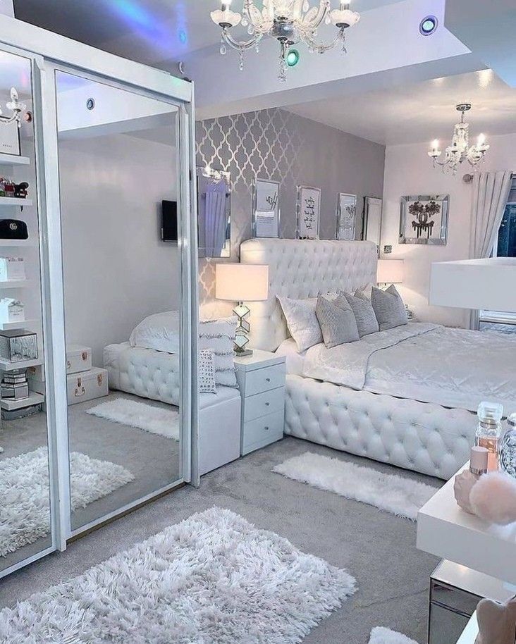Elegant modern luxury bedroom featuring a tufted white bed, crystal chandeliers, mirrored furniture, soft neutral décor, plush rugs, and sophisticated ambient lighting